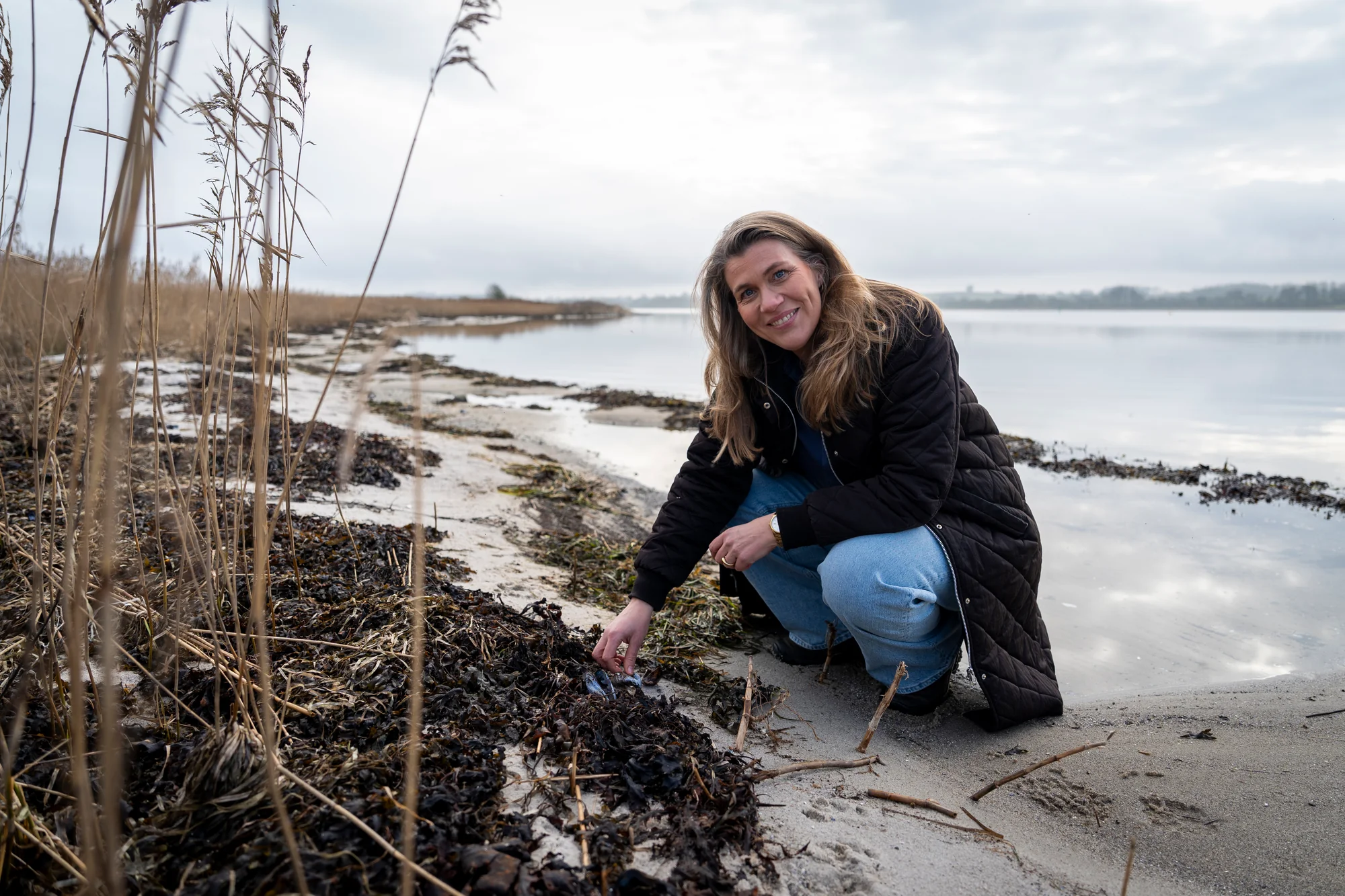 Mette Raun Hollesen, grundlægger af Kystnær, sidder på stranden ved kysten omgivet af tang og rør, iført sort jakke og jeans
